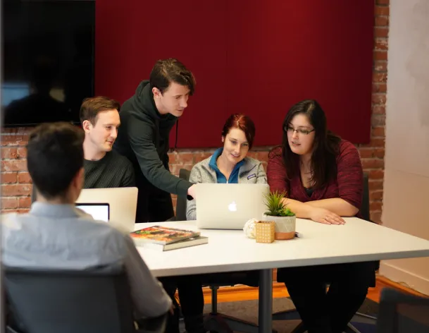 foto.umbii.boo team gathered around a laptop during a collaborative team meeting inside a creative office with brick walls and red acoustic panels.